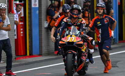 MotoGP rider in full gear on an orange KTM bike in the pit lane, flanked by crew in orange and navy.