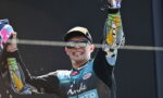 Motocross racer on a podium celebrating, arms raised in victory, wearing a blue/black racing suit, cap and sunglasses, with a cigarette in his mouth and sponsor logos visible.
