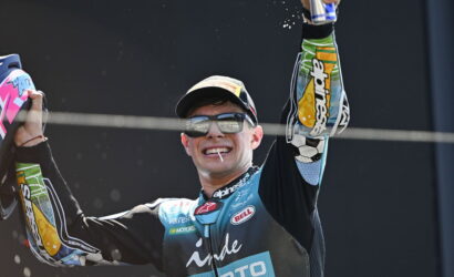Motocross racer on a podium celebrating, arms raised in victory, wearing a blue/black racing suit, cap and sunglasses, with a cigarette in his mouth and sponsor logos visible.
