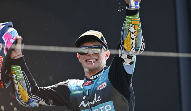 Motocross racer on a podium celebrating, arms raised in victory, wearing a blue/black racing suit, cap and sunglasses, with a cigarette in his mouth and sponsor logos visible.