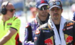 Moto racer in KTM Red Bull livery, cap and sunglasses, towel draped over shoulders, looking down.