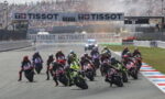 Motorcycle race start with a dense group of riders leaning into a turn on a track, crowds in stands and TISSOT banners overhead.