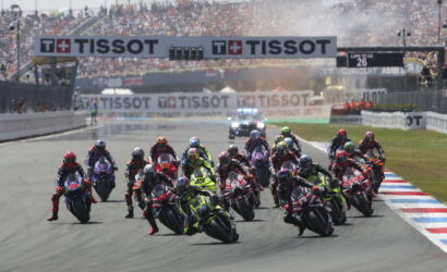 Motorcycle race start with a dense group of riders leaning into a turn on a track, crowds in stands and TISSOT banners overhead.