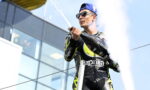 Motorsport rider in a black neon racing suit sprays champagne on a podium, wearing a blue cap and sunglasses.