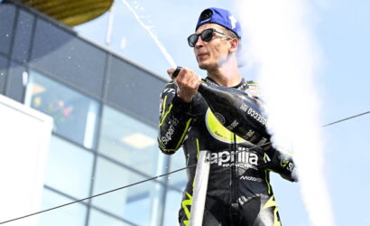 Motorsport rider in a black neon racing suit sprays champagne on a podium, wearing a blue cap and sunglasses.