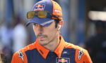 Man in orange and navy Red Bull KTM racing gear, wearing a cap with sunglasses perched on the brim and a wireless earbud in his ear, looking downward.”,