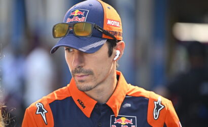Man in orange and navy Red Bull KTM racing gear, wearing a cap with sunglasses perched on the brim and a wireless earbud in his ear, looking downward.”,