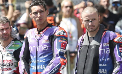 Two motorcyclists in purple Pramac racing suits stand with serious expressions in front of cheering spectators behind them.