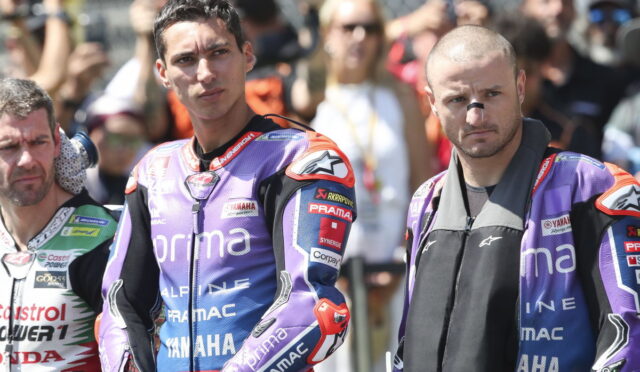 Two motorcyclists in purple Pramac racing suits stand with serious expressions in front of cheering spectators behind them.