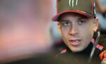 Close-up of a man wearing a black Monster cap and racing jacket, looking to the side during a conversation.
