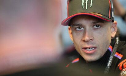 Close-up of a man wearing a black Monster cap and racing jacket, looking to the side during a conversation.