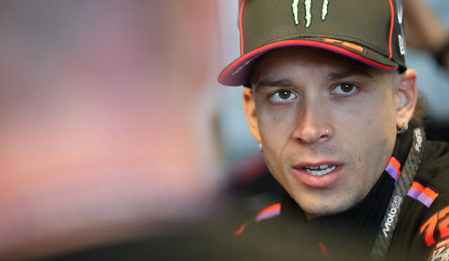 Close-up of a man wearing a black Monster cap and racing jacket, looking to the side during a conversation.