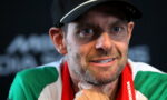 Close-up of a man with a cap and a green, white, and red scarf, looking slightly off to the side with a thoughtful smile.