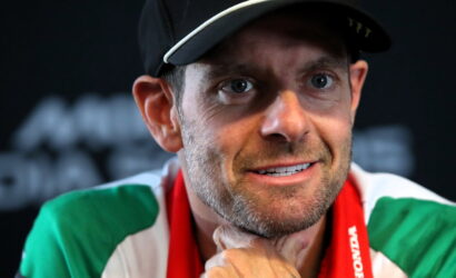 Close-up of a man with a cap and a green, white, and red scarf, looking slightly off to the side with a thoughtful smile.