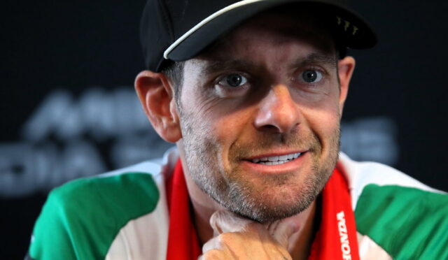 Close-up of a man with a cap and a green, white, and red scarf, looking slightly off to the side with a thoughtful smile.
