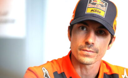 Close-up of a man wearing a Red Bull KTM cap and orange racing suit, looking slightly to the right.
