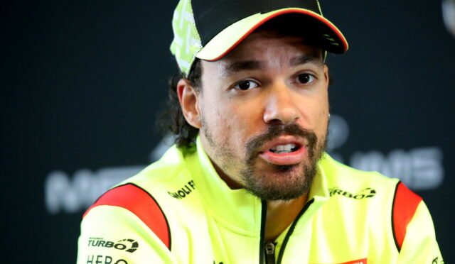 Close-up of a man in a neon yellow racing jacket and cap, speaking at a press conference.