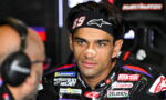 Close-up of a motor racing rider in a black headband with the number 89, a beige nose bandage, and a colorful racing suit, looking focused in the pit area