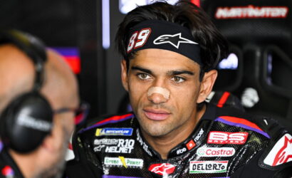 Close-up of a motor racing rider in a black headband with the number 89, a beige nose bandage, and a colorful racing suit, looking focused in the pit area