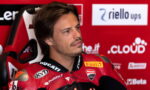 Man in a red Ducati racing suit sits in a red sponsor wall, looking off to the side with a thoughtful expression.