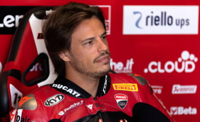 Man in a red Ducati racing suit sits in a red sponsor wall, looking off to the side with a thoughtful expression.