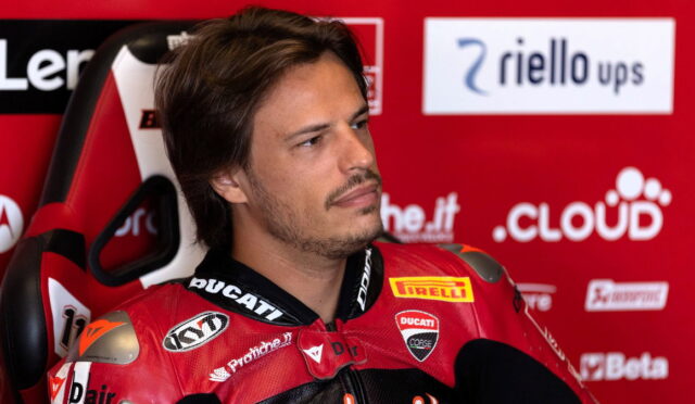 Man in a red Ducati racing suit sits in a red sponsor wall, looking off to the side with a thoughtful expression.