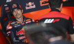 Rider in a KTM/Red Bull racing suit sits in a team chair, looking toward a crew member in a garage of sponsor logos.