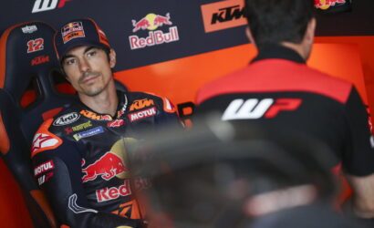 Rider in a KTM/Red Bull racing suit sits in a team chair, looking toward a crew member in a garage of sponsor logos.