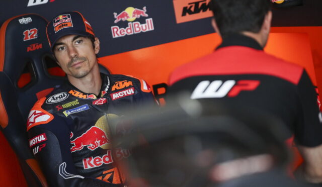 Rider in a KTM/Red Bull racing suit sits in a team chair, looking toward a crew member in a garage of sponsor logos.