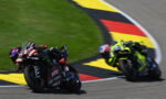 Two riders in a motorcycle race lean into a tight corner on a track with colorful curbing and grass nearby.