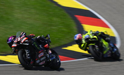 Two riders in a motorcycle race lean into a tight corner on a track with colorful curbing and grass nearby.