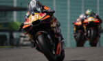 Motorcycle racer leaning into a turn on a race track, number 87 with Red Bull livery in focus; other rider in background