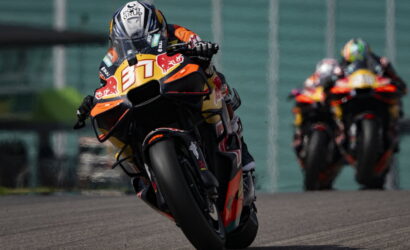 Motorcycle racer leaning into a turn on a race track, number 87 with Red Bull livery in focus; other rider in background
