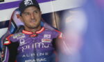 Moto racer in a purple Alpine?Pramac suit, seated in a racing cockpit, looking to the side with a cap on.