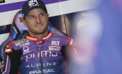 Moto racer in a purple Alpine?Pramac suit, seated in a racing cockpit, looking to the side with a cap on.
