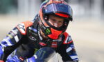 Motorcycle racer in a blue-black Yamaha suit wearing a red-orange helmet, leaning into a turn on a track.