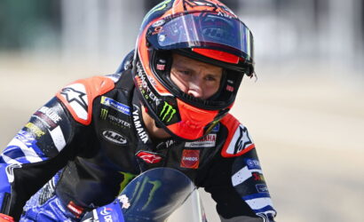 Motorcycle racer in a blue-black Yamaha suit wearing a red-orange helmet, leaning into a turn on a track.