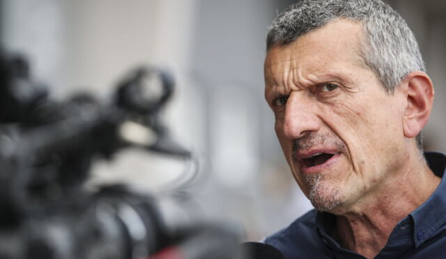 Man with gray hair speaks into a microphone during an interview, camera blurred in the foreground