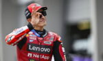 MotoGP rider in red Lenovo/NetApp suit makes an OK sign with his right hand, wearing a red cap and sunglasses.