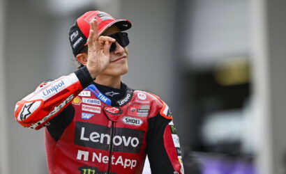MotoGP rider in red Lenovo/NetApp suit makes an OK sign with his right hand, wearing a red cap and sunglasses.