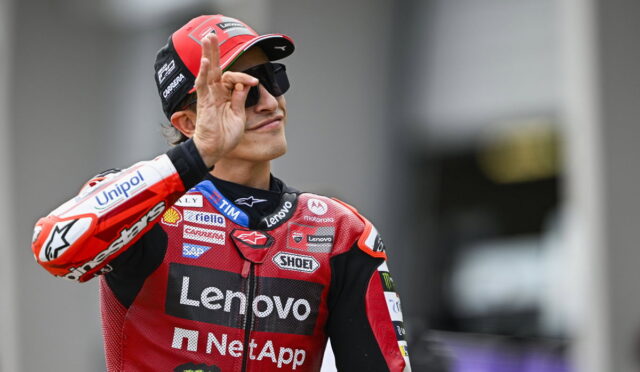 MotoGP rider in red Lenovo/NetApp suit makes an OK sign with his right hand, wearing a red cap and sunglasses.