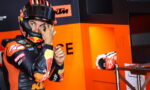 Moto racer in orange-and-black gear adjusts his helmet while standing in a bright orange pit area
