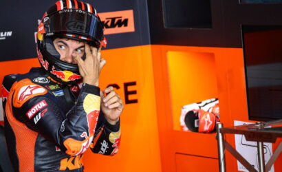 Moto racer in orange-and-black gear adjusts his helmet while standing in a bright orange pit area