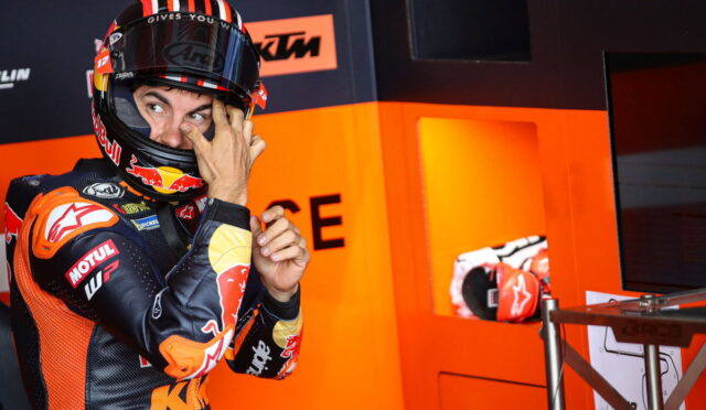 Moto racer in orange-and-black gear adjusts his helmet while standing in a bright orange pit area