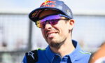 Portrait of a male race car driver smiling, wearing a blue racing shirt, a Red Bull cap, and mirrored sunglasses.