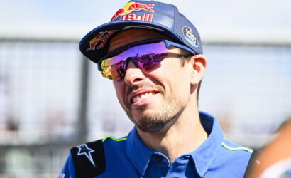 Portrait of a male race car driver smiling, wearing a blue racing shirt, a Red Bull cap, and mirrored sunglasses.