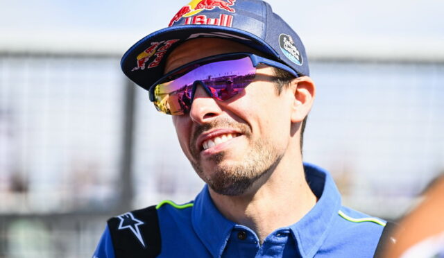 Portrait of a male race car driver smiling, wearing a blue racing shirt, a Red Bull cap, and mirrored sunglasses.