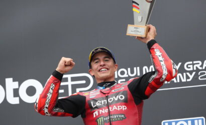MotoGP rider in red sponsor suit celebrates on podium, raising a trophy above his head with a wide smile.