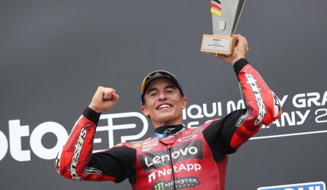 MotoGP rider in red sponsor suit celebrates on podium, raising a trophy above his head with a wide smile.