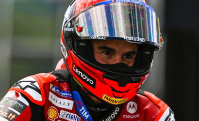 Close-up of a motor racing driver's red helmet and suit, mirrored visor showing focus, with sponsor logos like Lenovo and Parmacotto visible on the gear